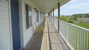 A covered porch with wooden flooring and white railings, overlooking a grassy area and a body of water. - Coho Motel and Suites (Kewaunee)