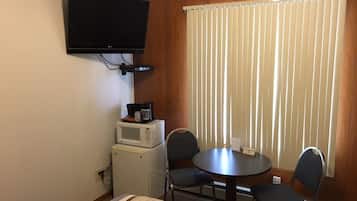 A hotel room with a bed, a TV mounted on the wall, a small refrigerator, a microwave, a table with two chairs, and a window with blinds.