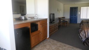 A compact kitchen with a refrigerator, microwave, and sink. There is a television on a wooden cabinet. A dining area with a table and chairs is visible, and a blue door is in the background. - Coho Motel and Suites (Kewaunee)