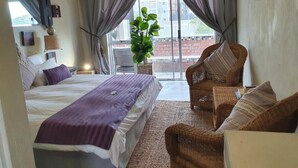 A bedroom with a bed, a wicker chair, a potted plant, and a view of the outside. - Melite Rooms (Cape Town)