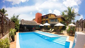 Una piscina con sillas de descanso y sombrillas, rodeada de edificios con temática tropical.