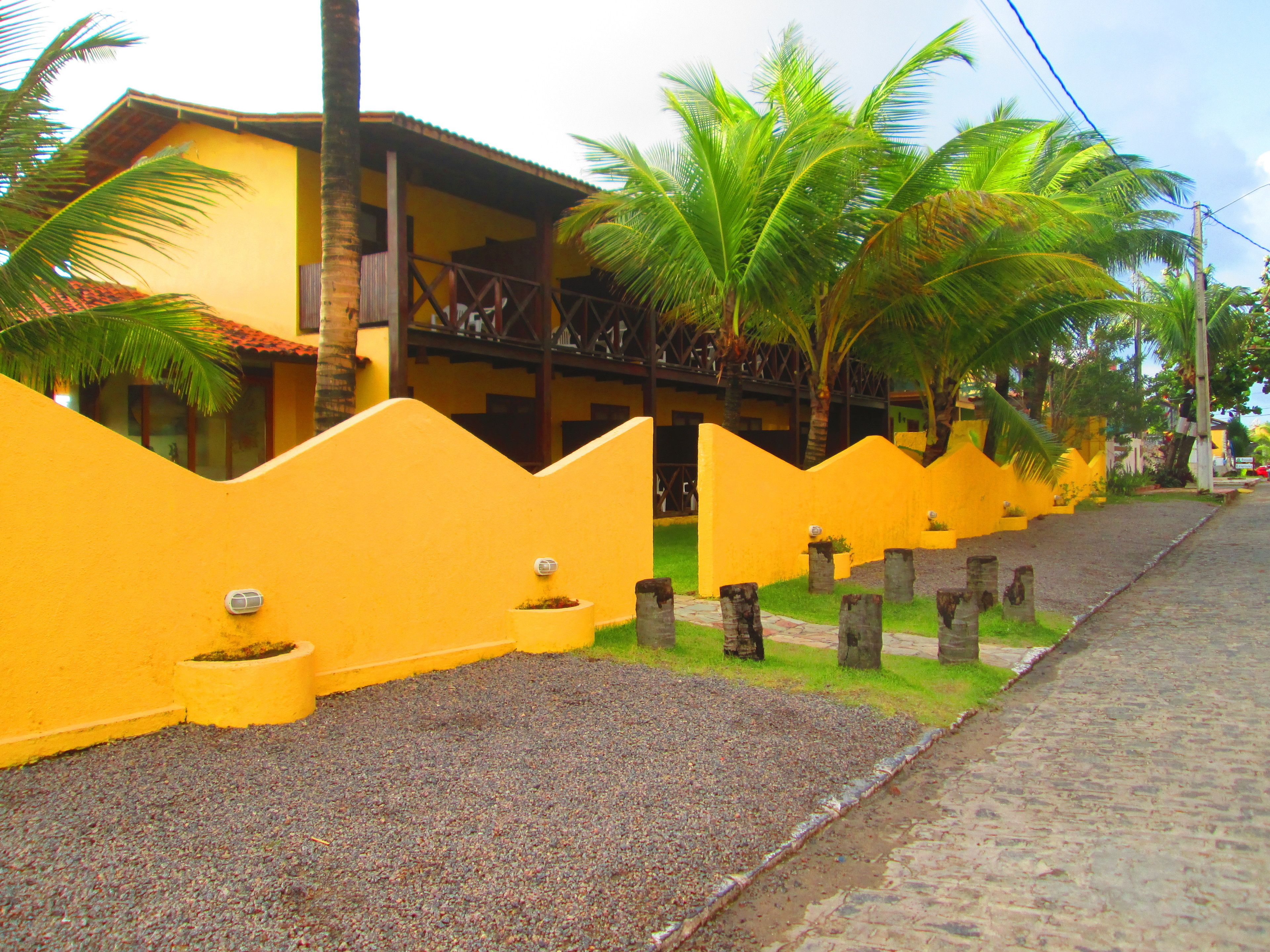 A yellow building with a balcony, surrounded by palm trees and a gravel path.