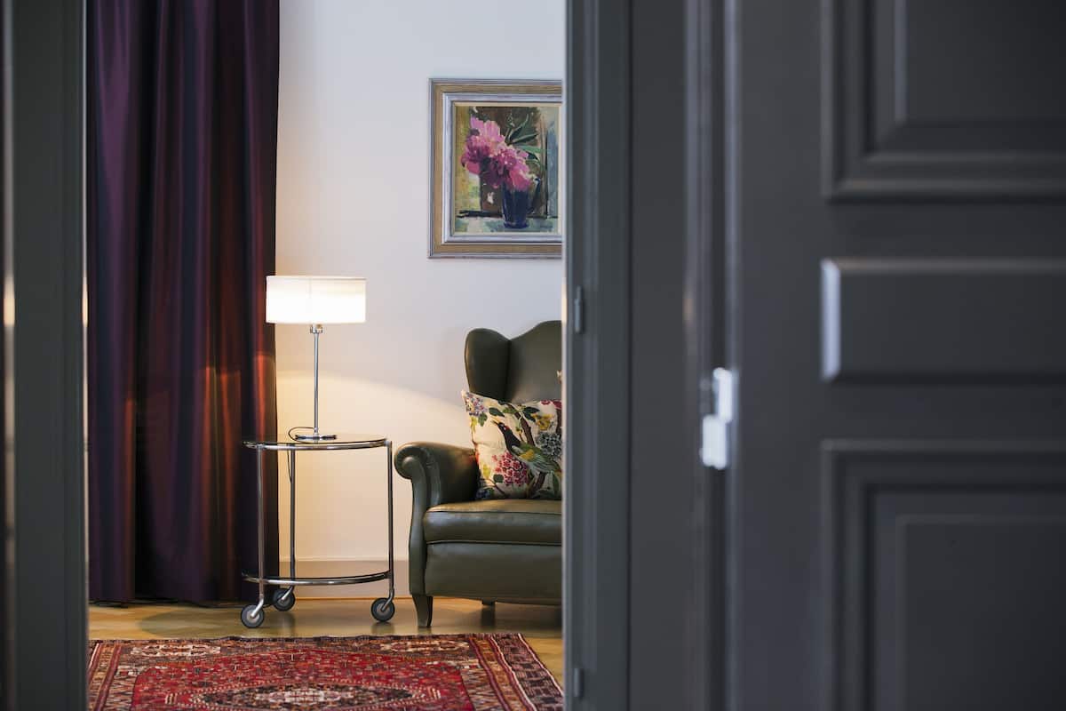 Una habitación con una puerta oscura, un sillón verde con un cojín floral, una mesita con una lámpara, un cuadro enmarcado en la pared y una alfombra roja con estampado.