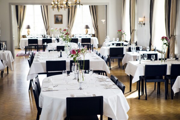 Un comedor con varias mesas dispuestas para una comida, con manteles blancos, sillas negras y centros de mesa florales.