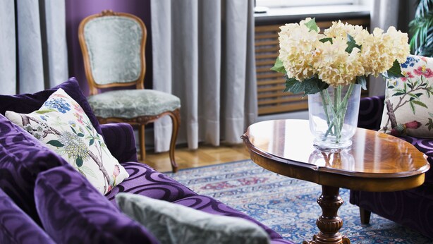 Un sofá morado con cojines estampados, una mesa redonda de madera con un jarrón de flores, y una silla tapizada de color verde en una habitación con alfombra y cortinas de diseño.