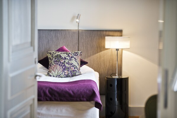 Una cama bien hecha con una colcha morada y una almohada con motivos florales, una lámpara de noche moderna y un cabecero de madera.