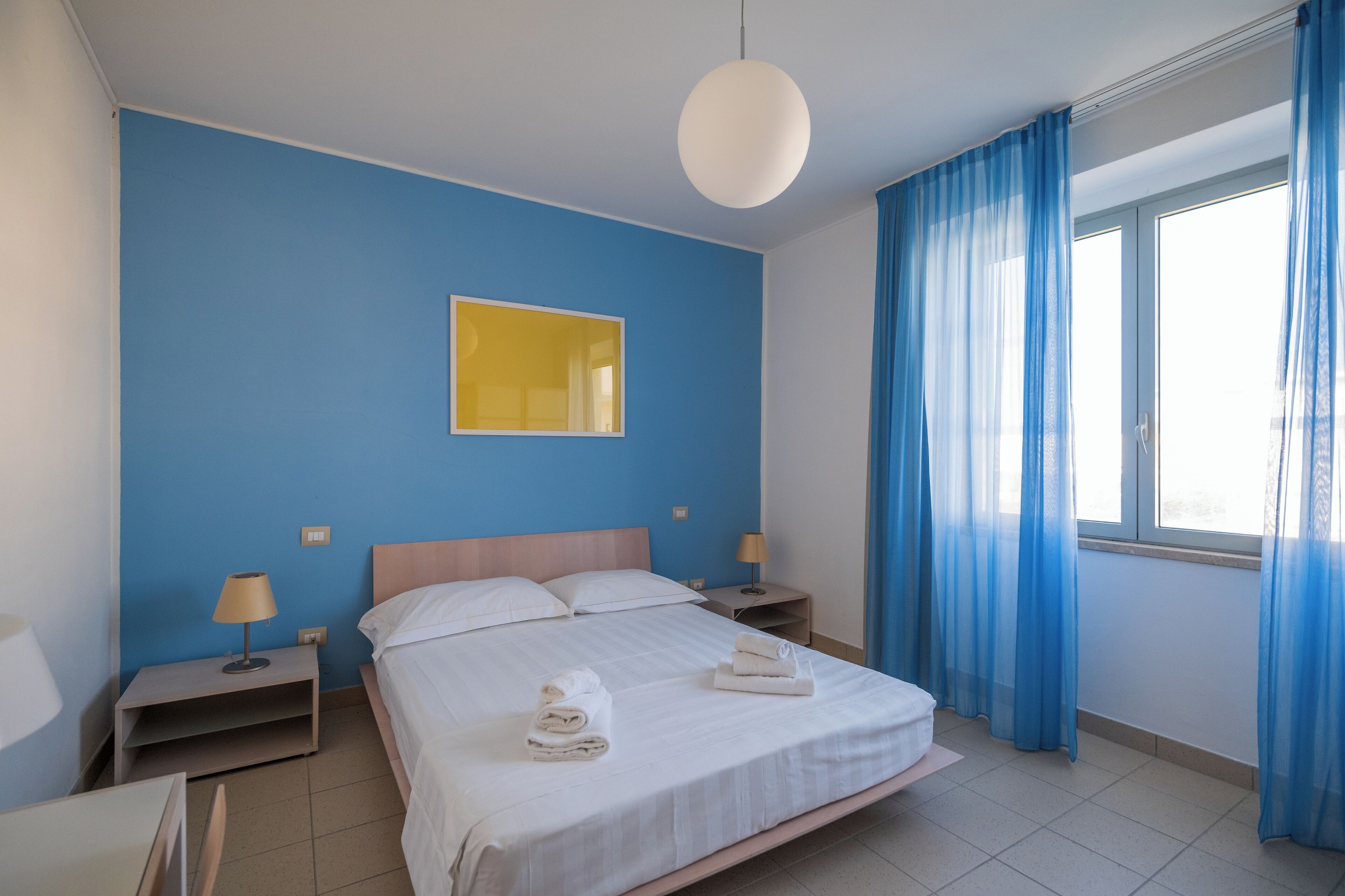 A neatly arranged hotel room with a large bed, two bedside tables, a bedside lamp, a wall-mounted mirror, and a window with blue curtains.
