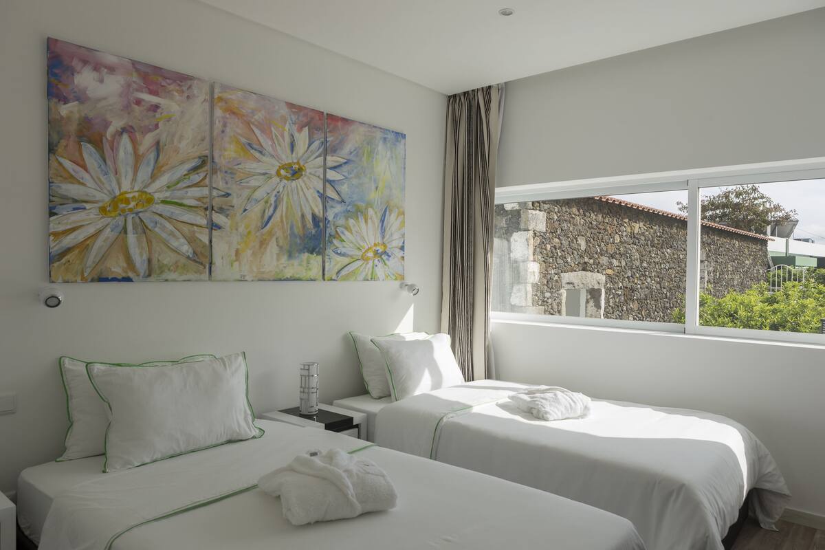A hotel room with two beds, a large floral painting, a stone wall, and a view of greenery outside.