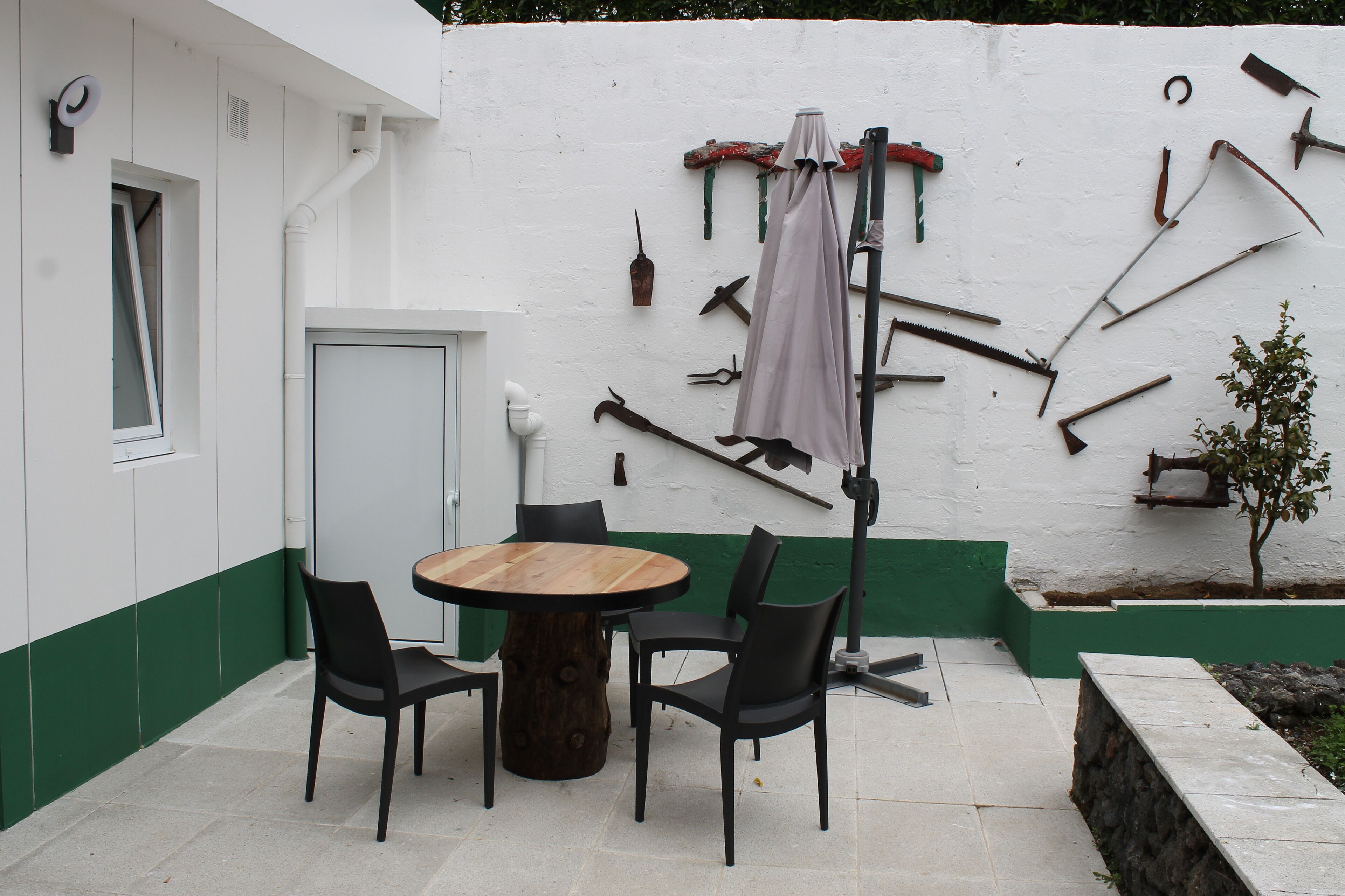 Un patio con una mesa de madera redonda y cuatro sillas, una pared blanca con detalles verdes y diversas herramientas colgadas.
