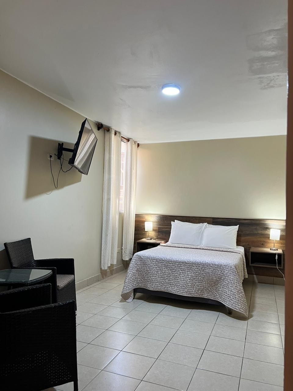Habitación de hotel con cama, televisor montado en la pared, mesa con sillas y mesita de noche con lámpara.
