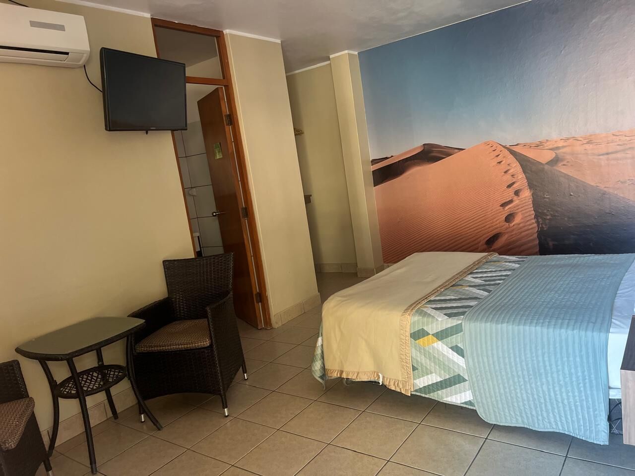 A hotel room with a bed, a small table, a chair, a television, and a desert mural on the wall.