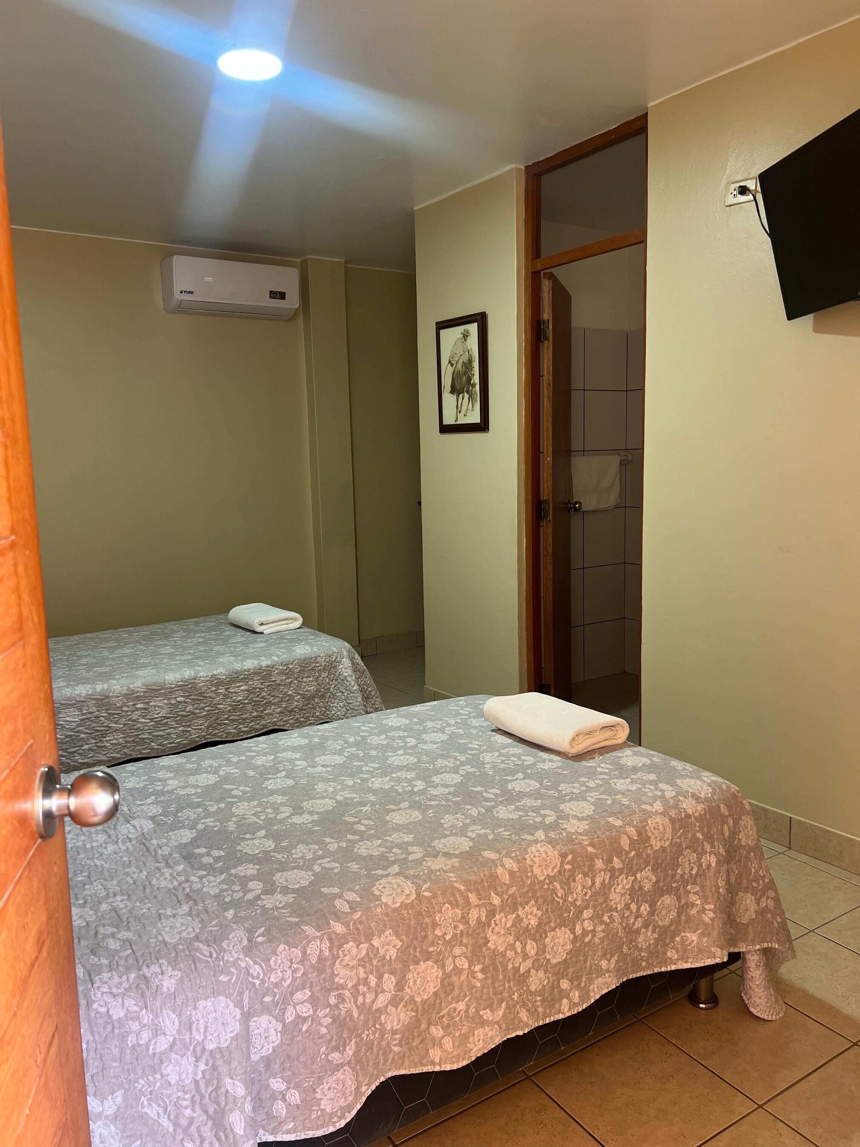 A hotel room with two beds, a mounted TV, and an air conditioning unit.