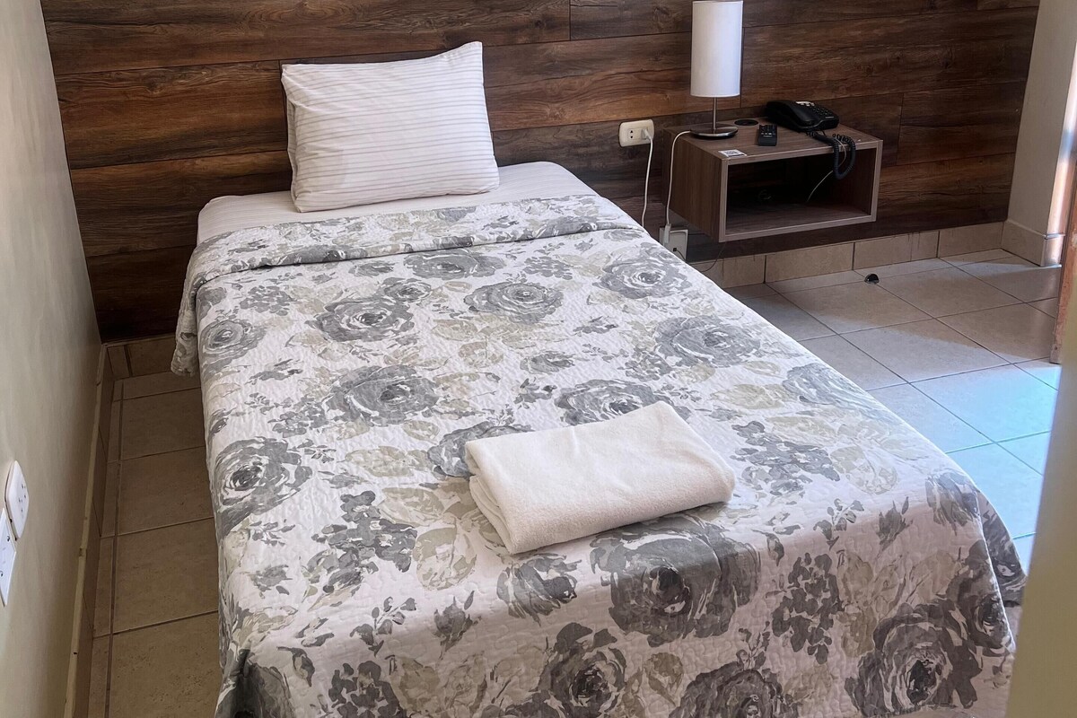 Una cama bien tendida con una colcha estampada, una lámpara de noche y una mesita de noche de madera con un teléfono y un cargador.
