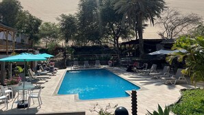 A swimming pool surrounded by lounge chairs and umbrellas, with palm trees and a desert landscape in the background.