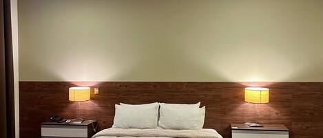 A hotel room with a large bed, two bedside tables with lamps, a wooden headboard, and a plain wall.