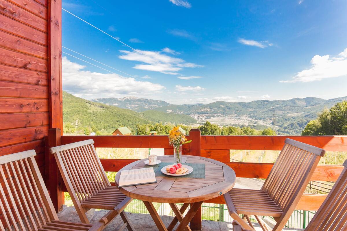 A wooden deck with two chairs and a table, overlooking a mountainous landscape.