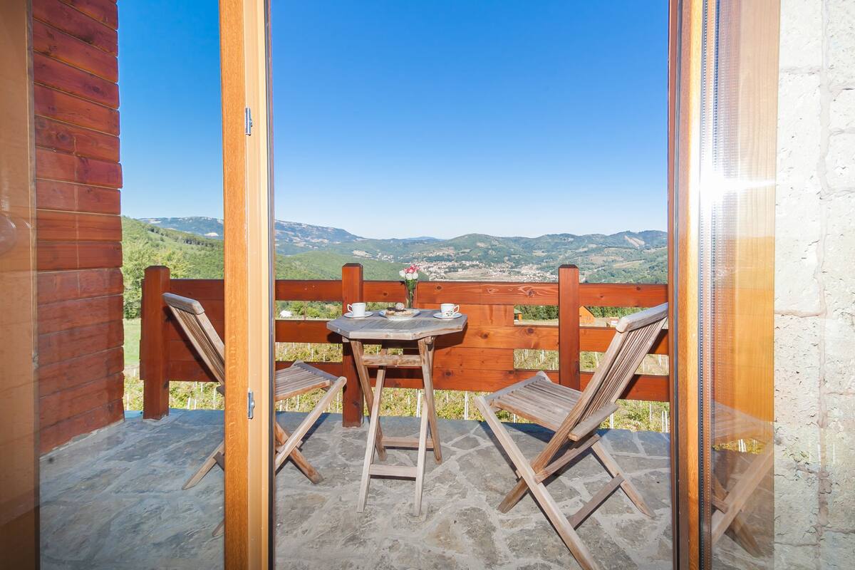 A wooden balcony with a table and two chairs, overlooking a scenic landscape.