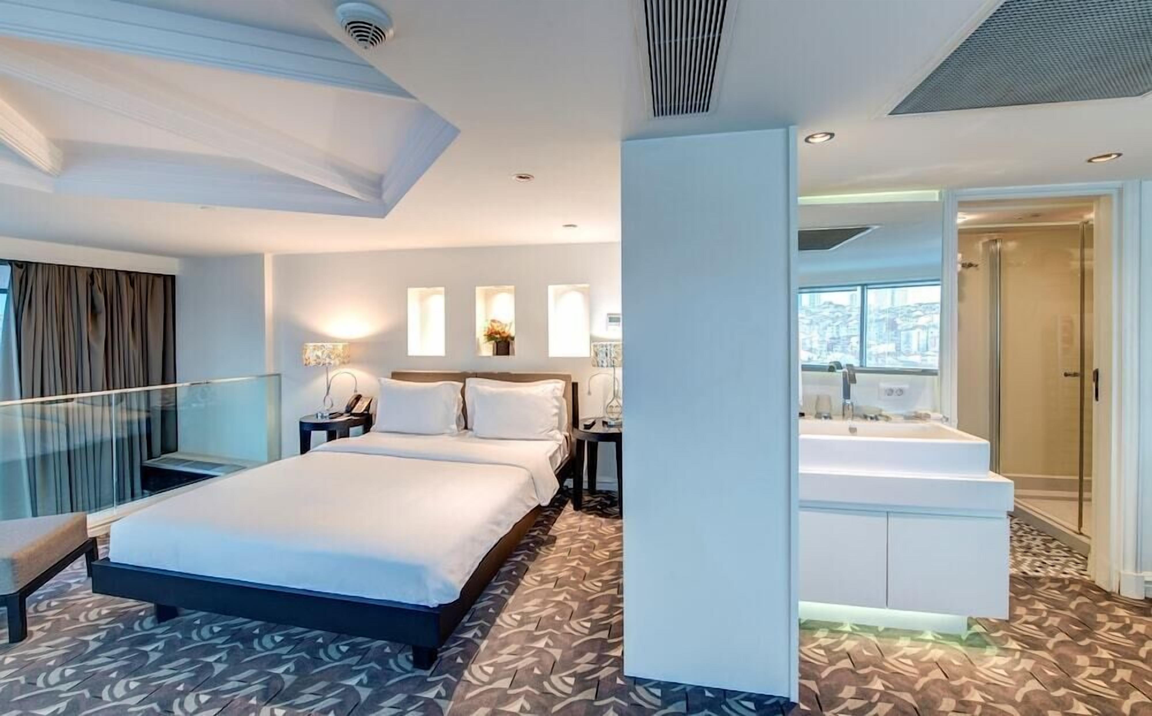 A hotel room with a large bed, a sitting area, a bathroom with a bathtub, and a view of the city.