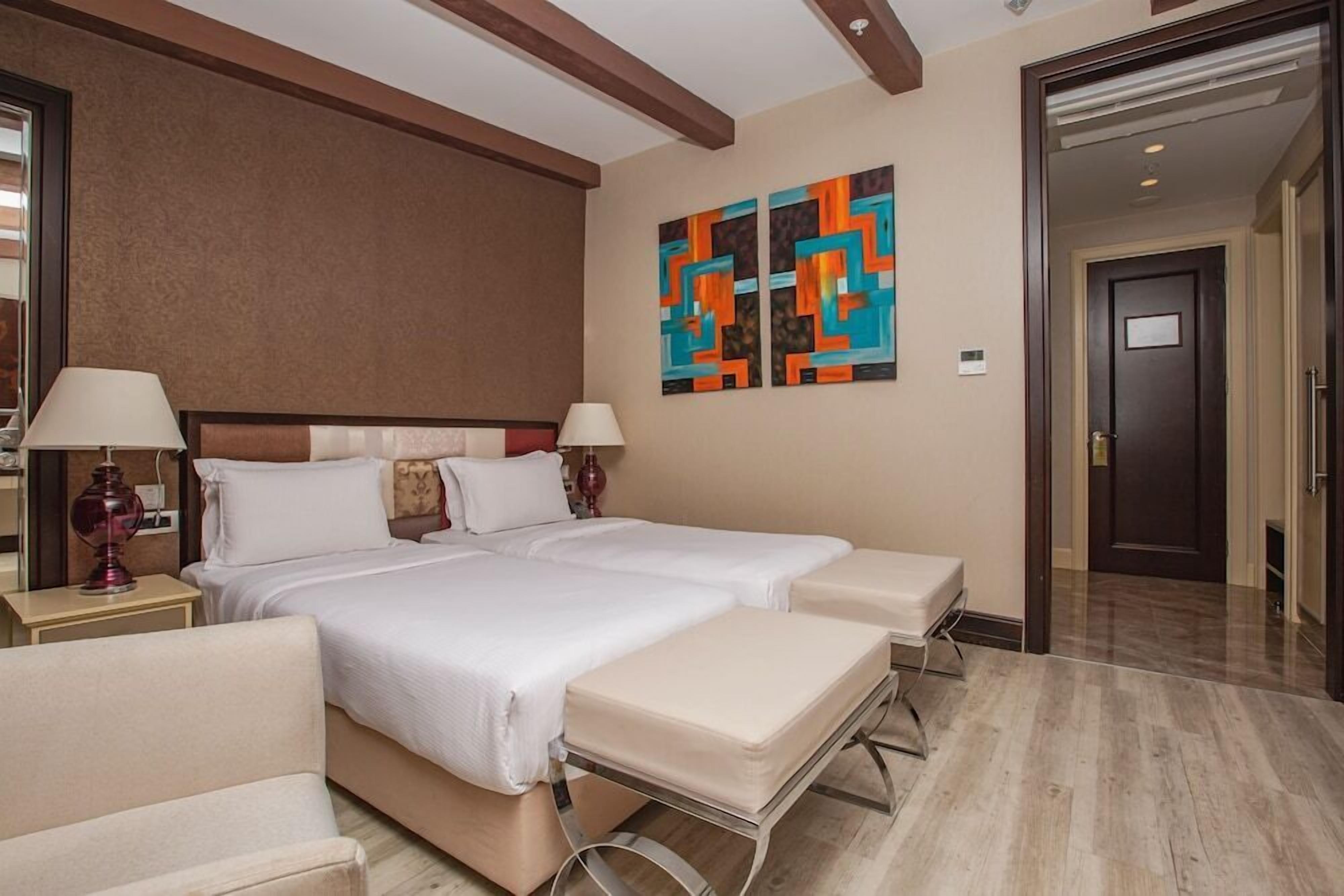 A hotel room with a double bed, two bedside tables with lamps, a bench, and a door leading to another room.