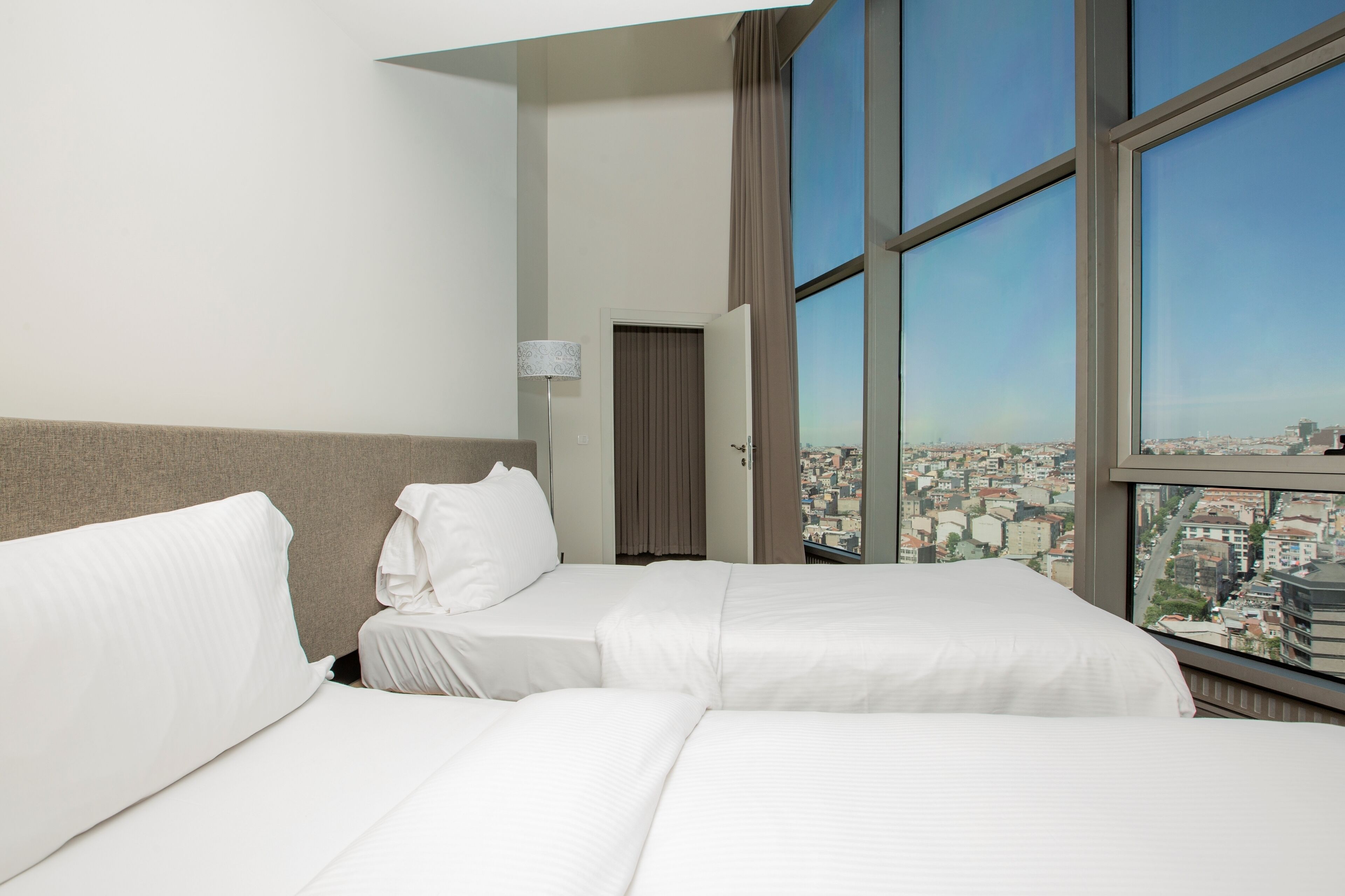 A hotel room with two beds, a large window with a city view, and a door leading to another room.
