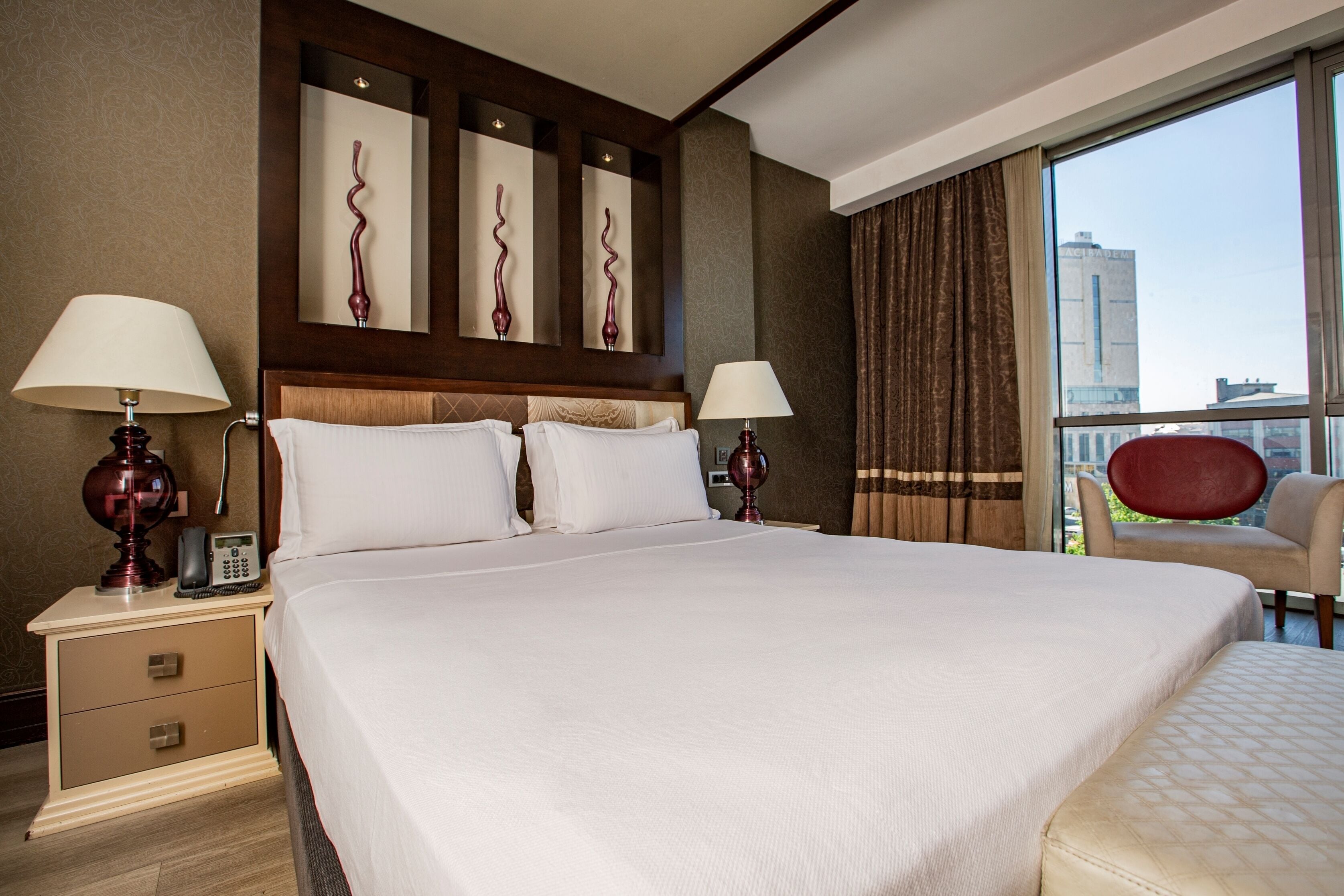 A hotel room with a large bed, two bedside tables with lamps, a mirror, and a view of a cityscape through a large window.