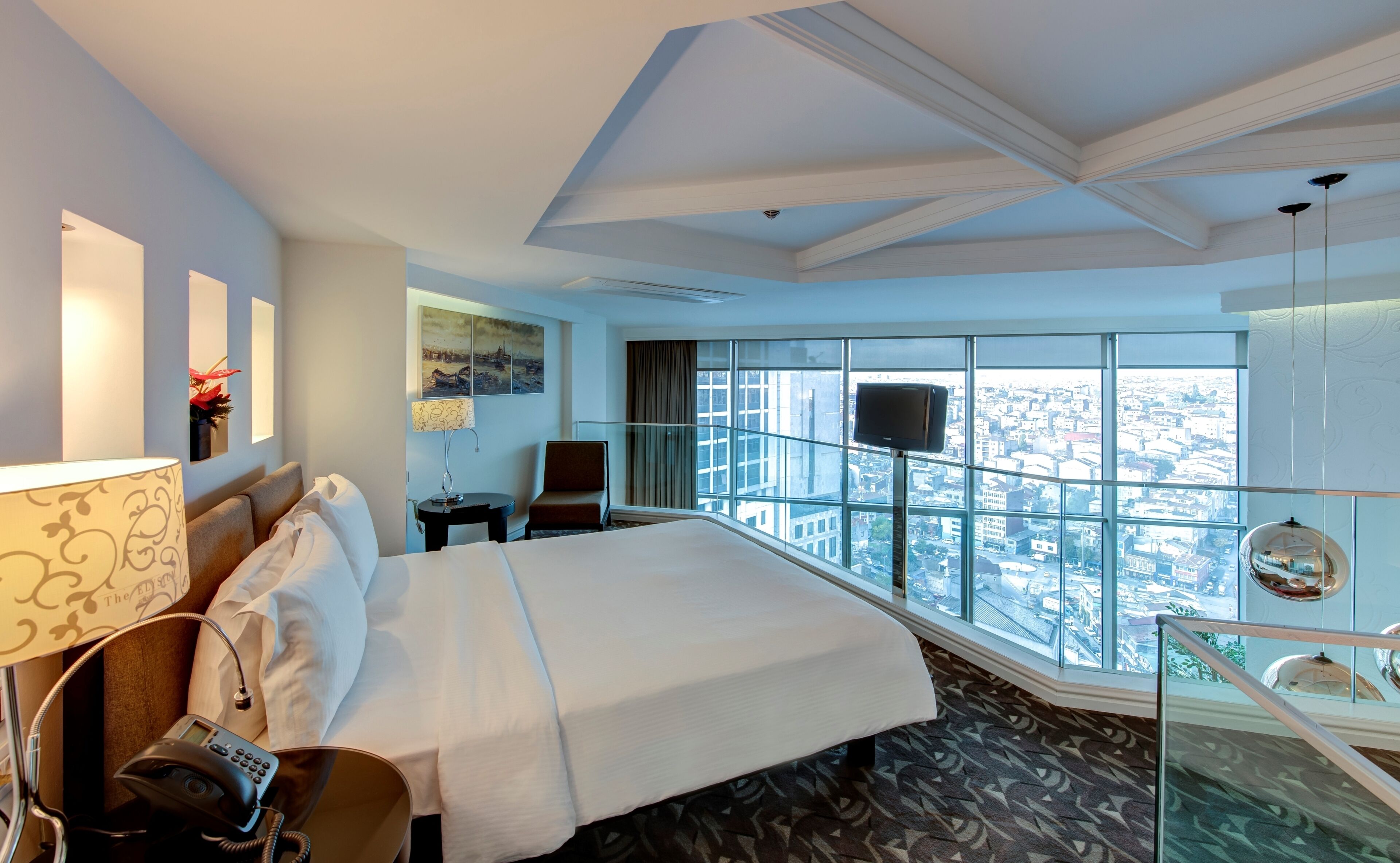 A hotel room with a large bed, a desk with a telephone, a chair, a small table with a lamp, a painting on the wall, and a large window offering a city view.