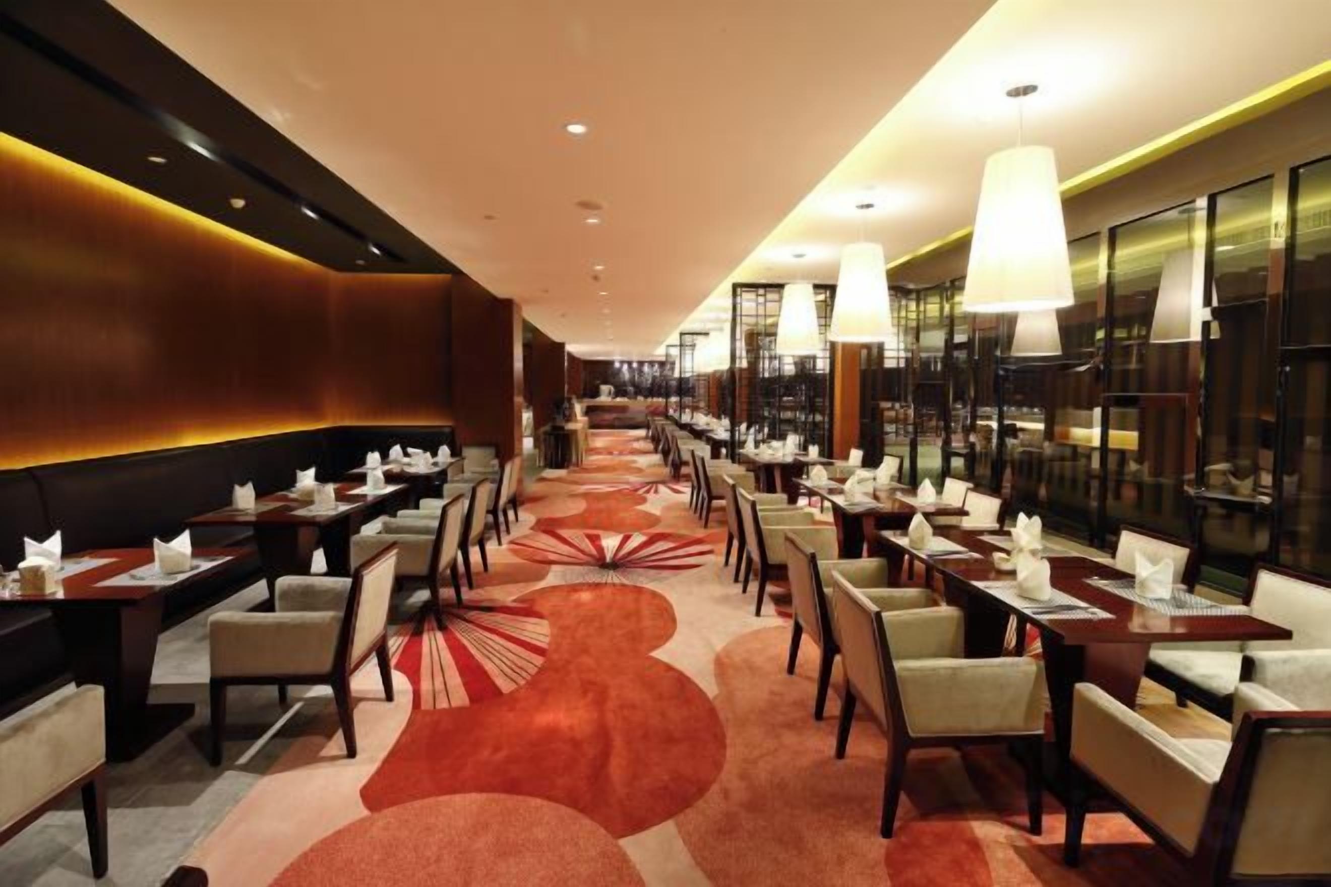 A hotel dining area with multiple tables and chairs, a patterned carpet, and modern lighting fixtures.