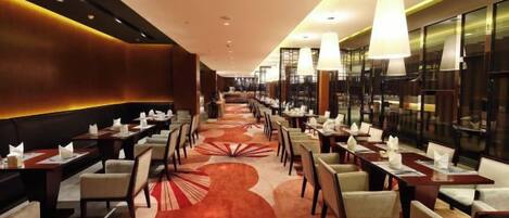 A hotel dining area with multiple tables and chairs, a patterned carpet, and modern lighting fixtures.