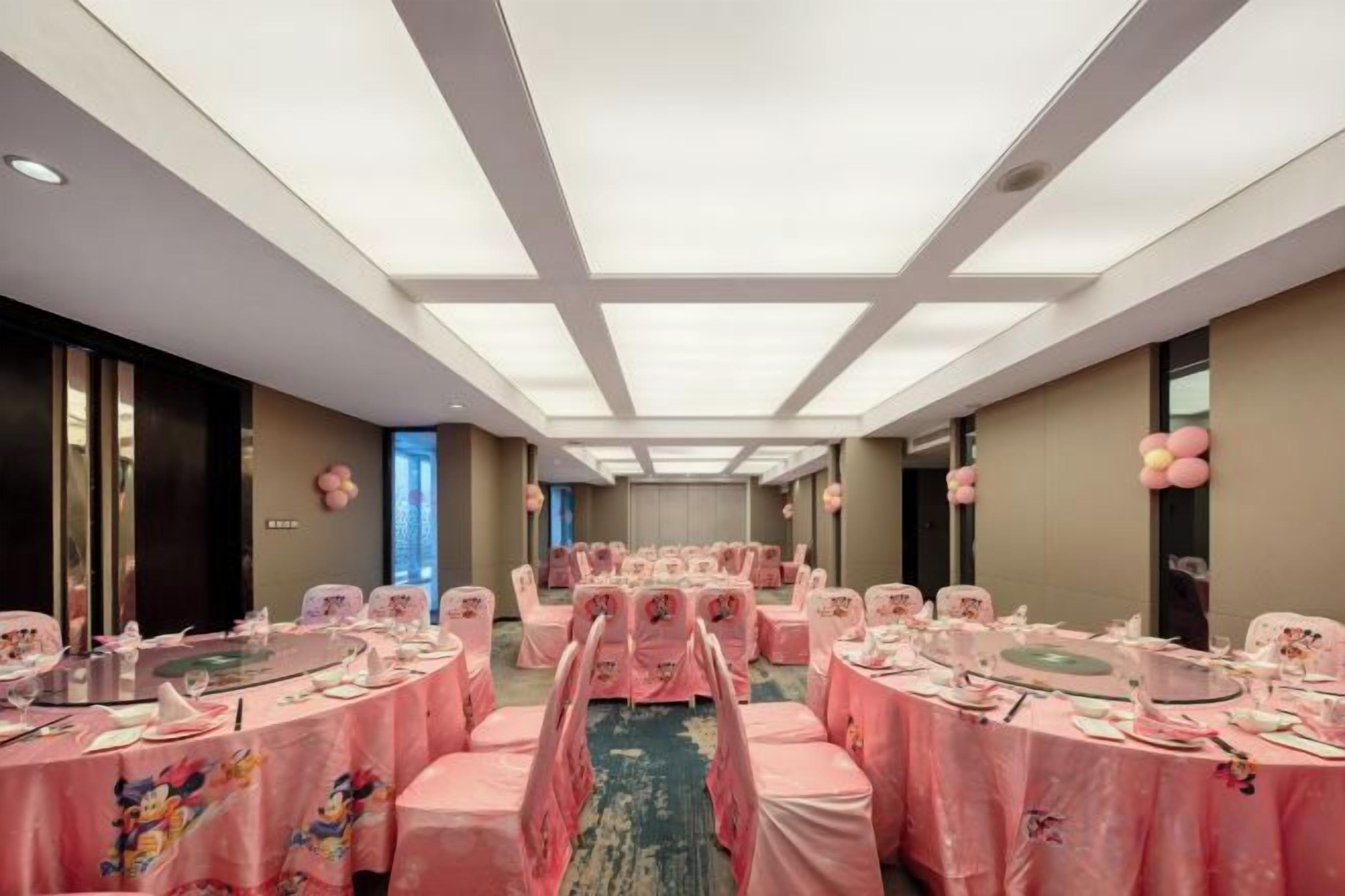 A hotel room with a long corridor, multiple round tables with pink tablecloths, and chairs.