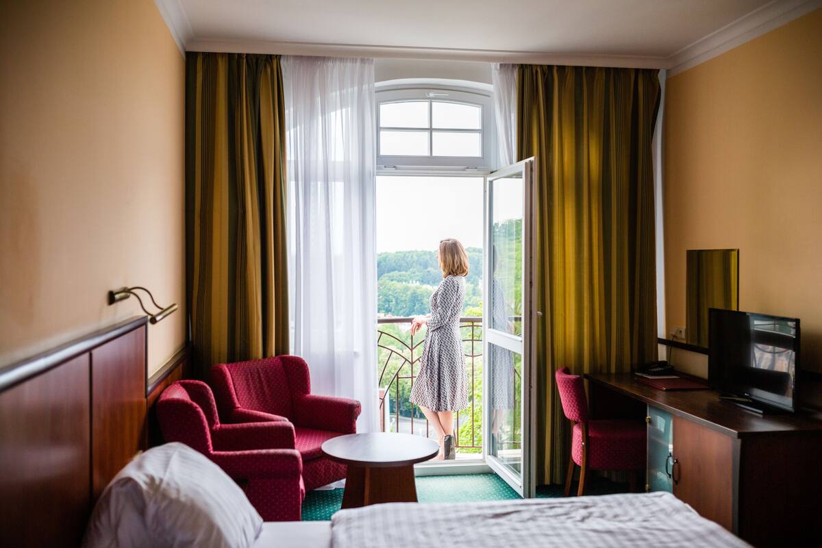 A hotel room with a bed, a desk with a television, a red chair, a small table, and a balcony with a view of greenery.