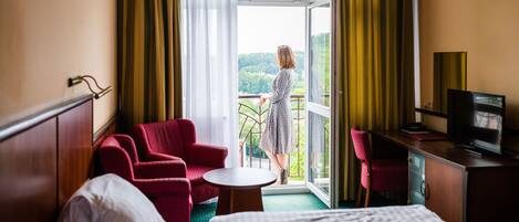 A hotel room with a bed, a desk with a television, a red chair, a small table, and a balcony with a view of greenery.