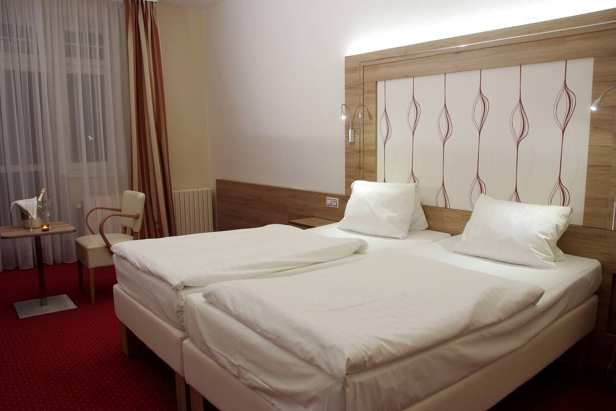 A hotel room with two beds, a wooden headboard, a small table with a bottle and glasses, and a chair.
