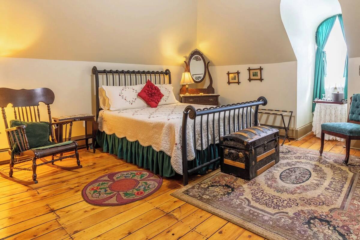 Un dormitorio con suelo de madera, una cama con cubrecama blanco y verde, una mecedora, un sillón azul y una maleta antigua.
