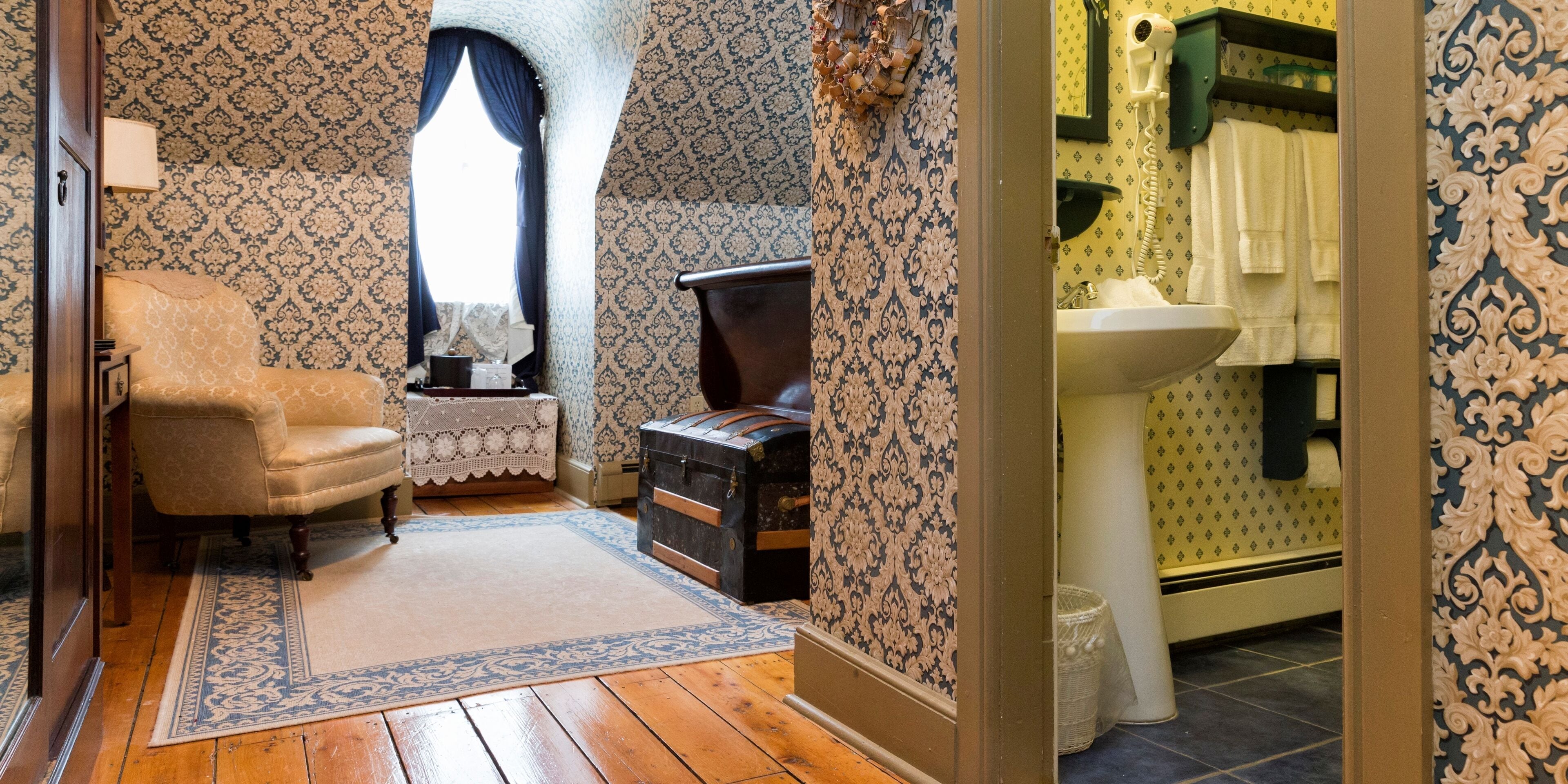 Una habitación de estilo antiguo con papel tapiz estampado, suelo de madera, una silla vintage, una pequeña mesa con mantel de encaje y un baúl.