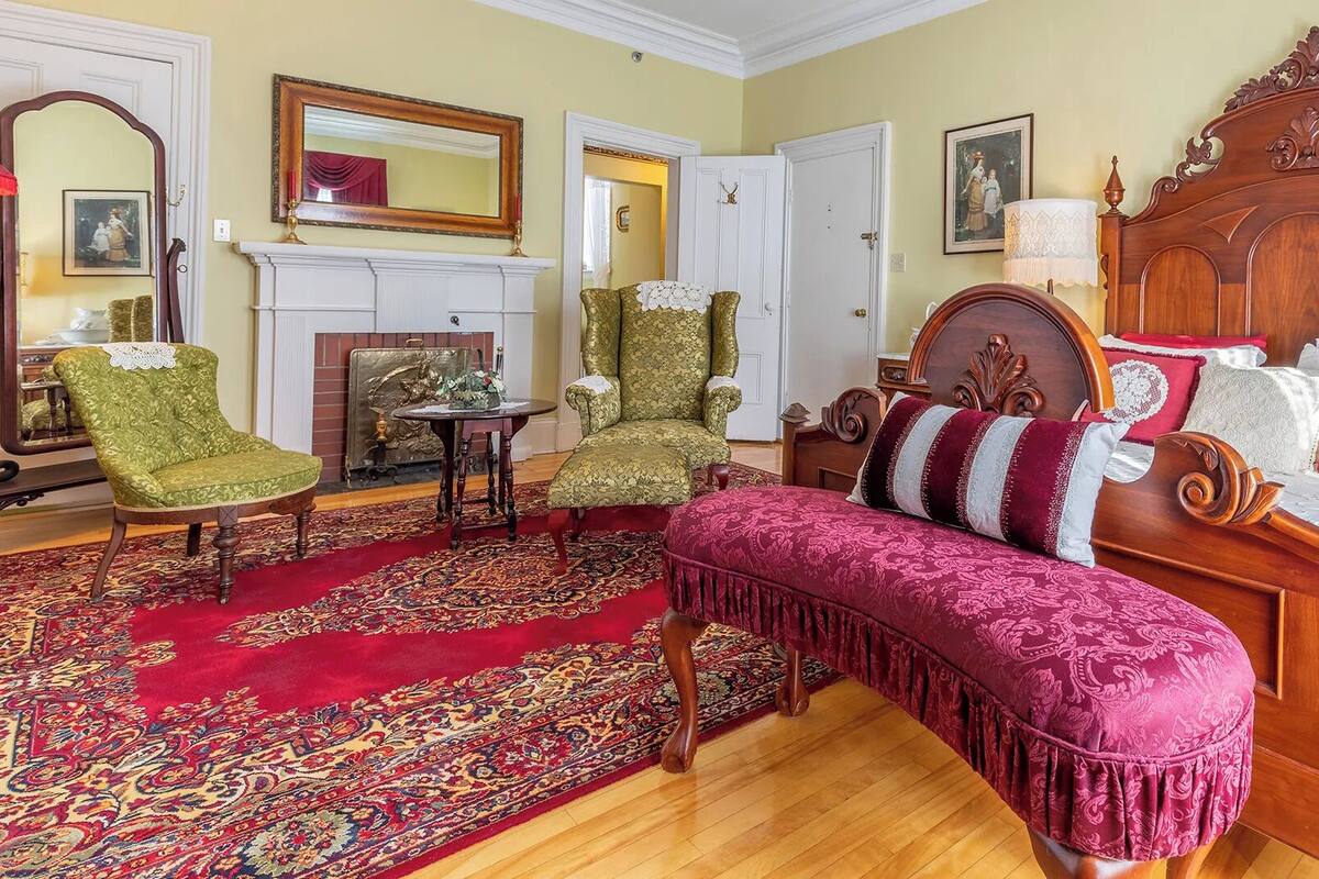 Una habitación tradicional con una cama de madera, sillones verdes, un tapete rojo estampado y una chimenea.