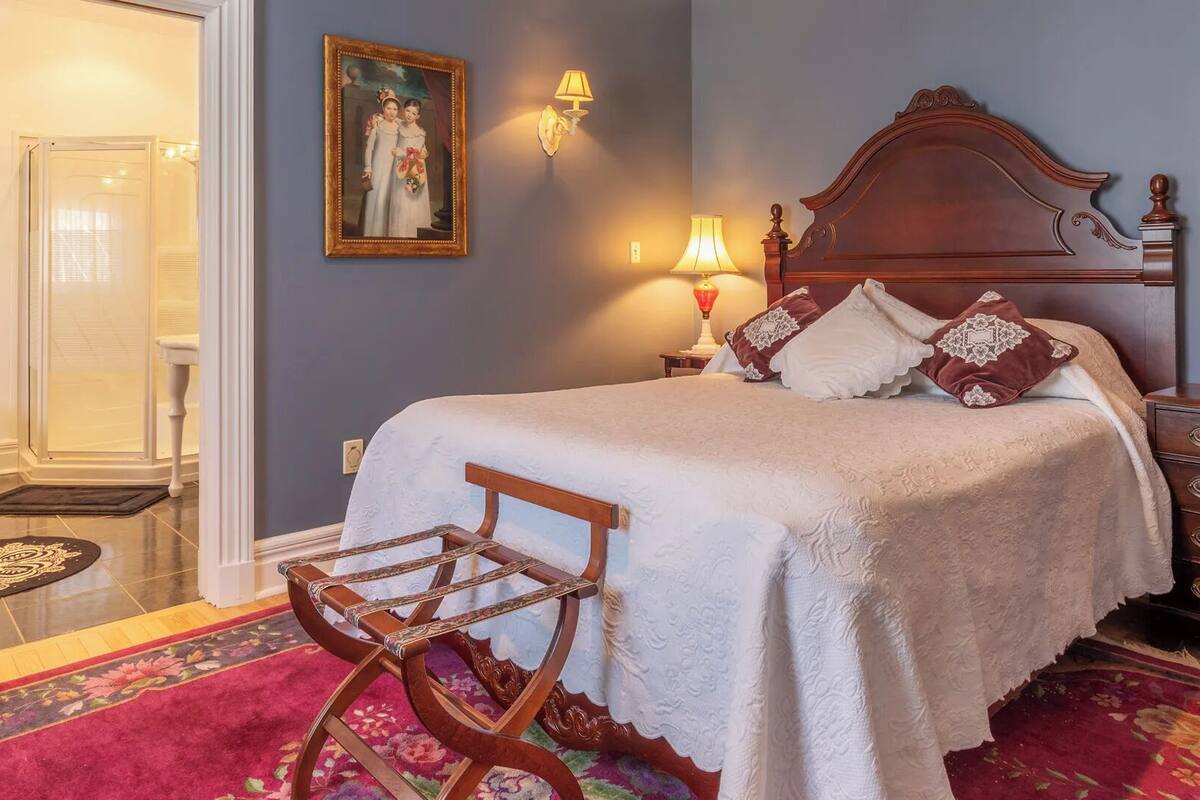 Un dormitorio con cama, una silla de madera, cómoda, espejo y un cuadro en la pared.
