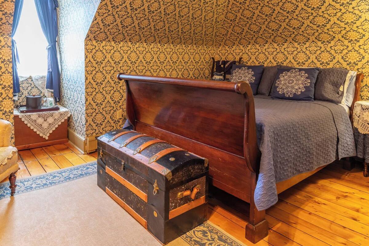 Un dormitorio tradicional con cama con dosel, un baúl antiguo y un sillón clásico.