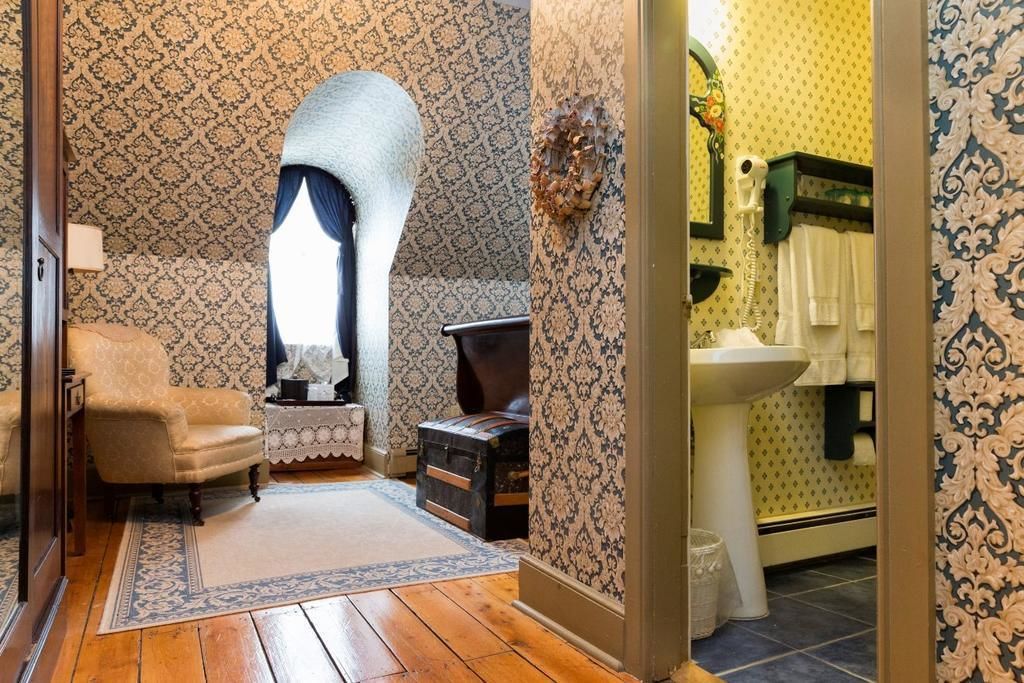 A room with patterned wallpaper, a wooden floor, a vintage armchair, a small table with a lamp, and a window with curtains.