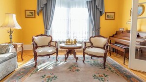 Una stanza con una parete gialla, un pianoforte, due poltrone decorate, un tavolo rotondo con un servizio da tè e un tappeto floreale.