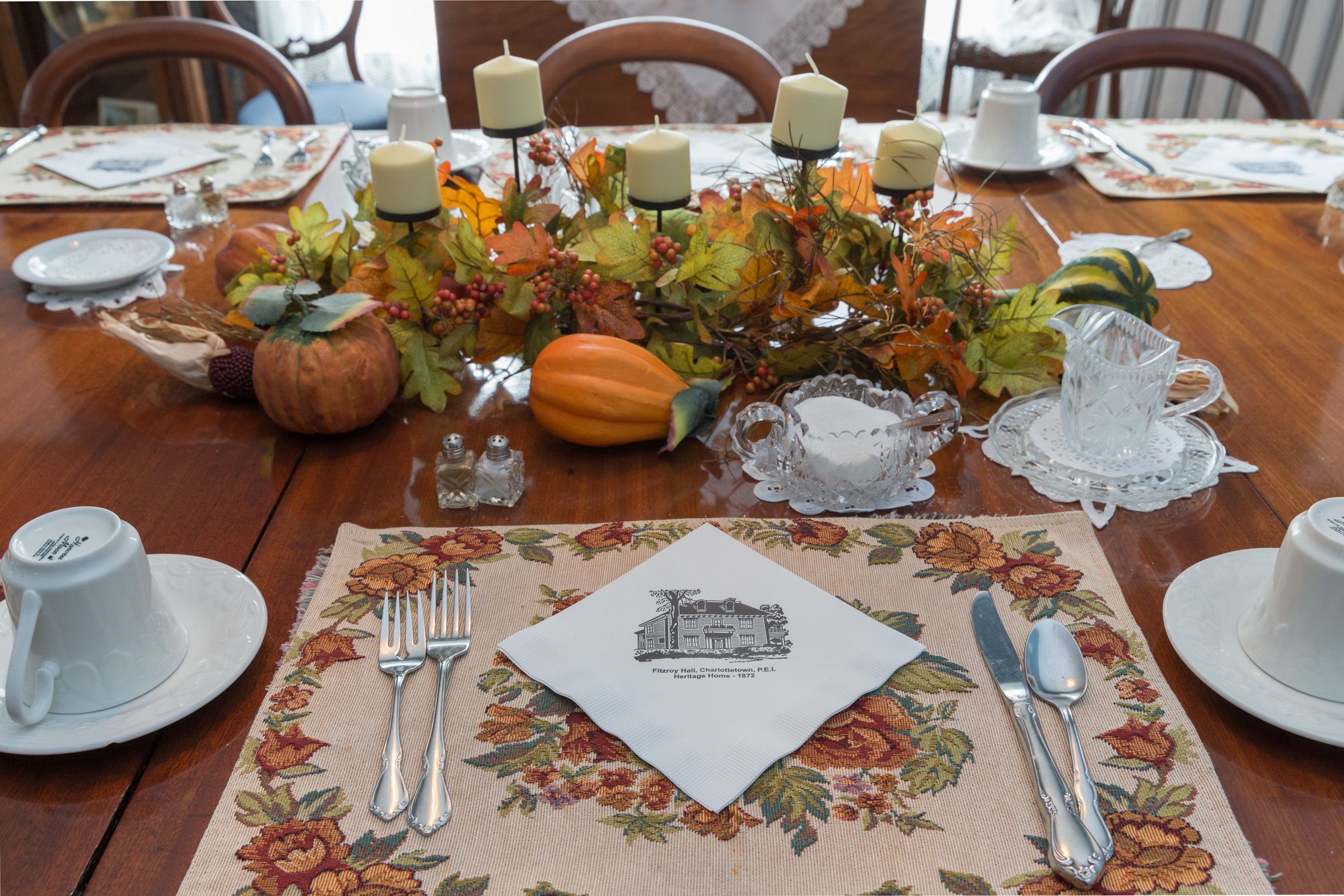 A dining table set with a floral tablecloth, a placemat with a building illustration, and autumn-themed decorations including pumpkins and candles.