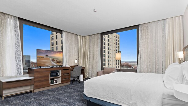 Quarto de hotel moderno com uma cama grande, uma escrivaninha com computador, uma cadeira e uma televisão.