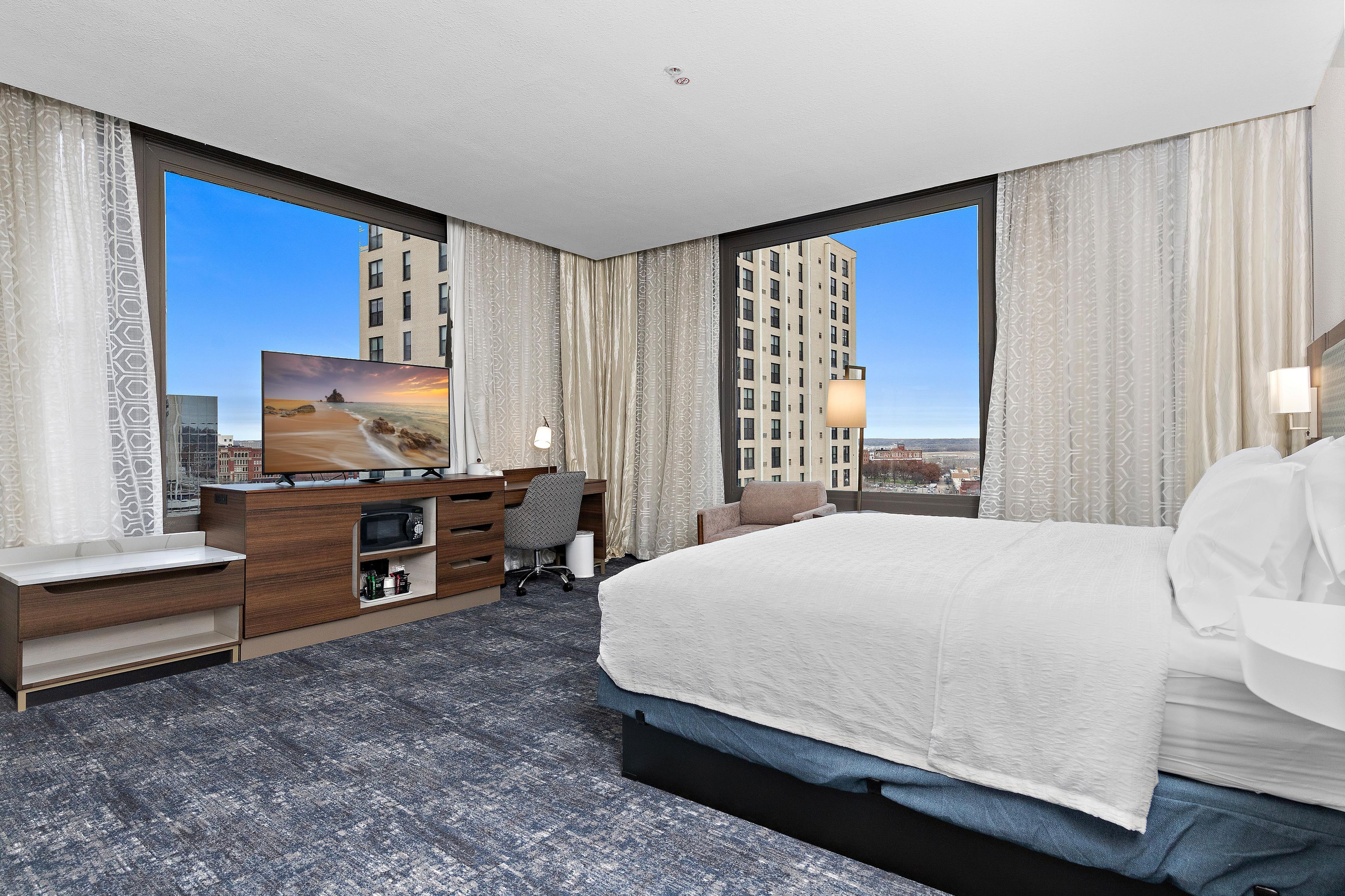 A hotel room with a large bed, a desk with a computer, a television, and a view of a cityscape.