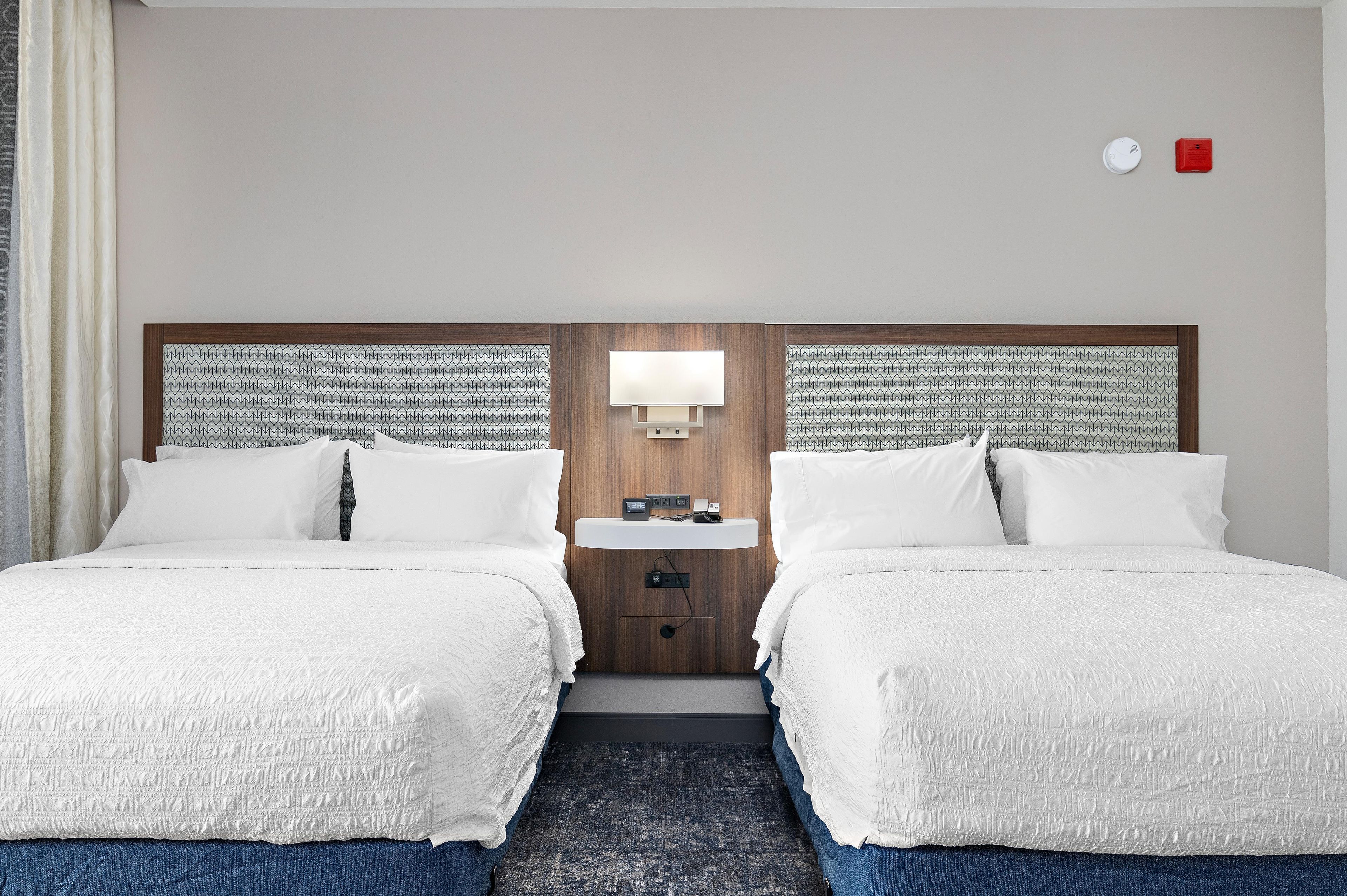 A hotel room with two beds, a bedside table with a lamp, and a small wall-mounted device.