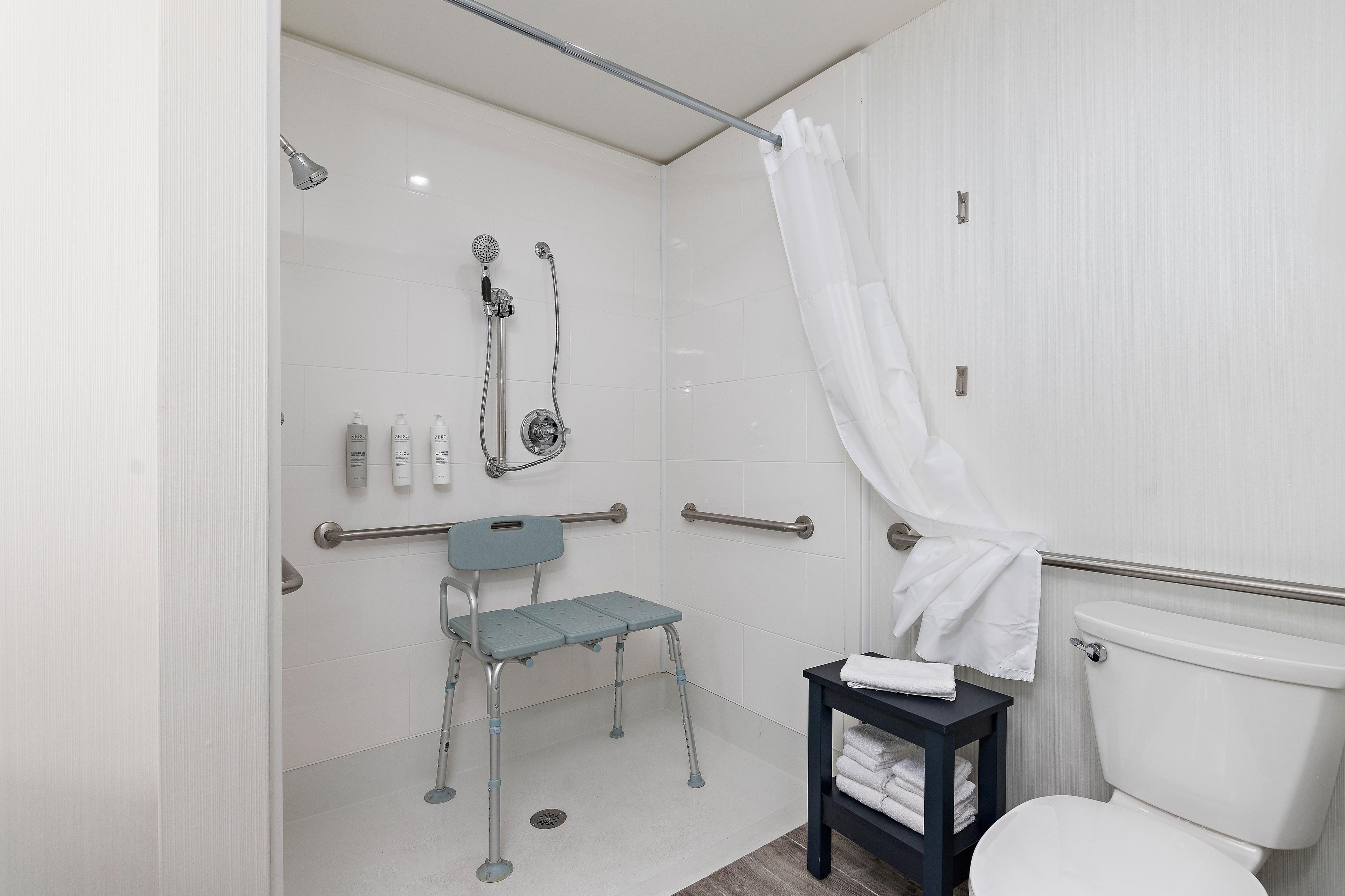 Un baño con ducha, inodoro, una mesita con toallas y una silla.