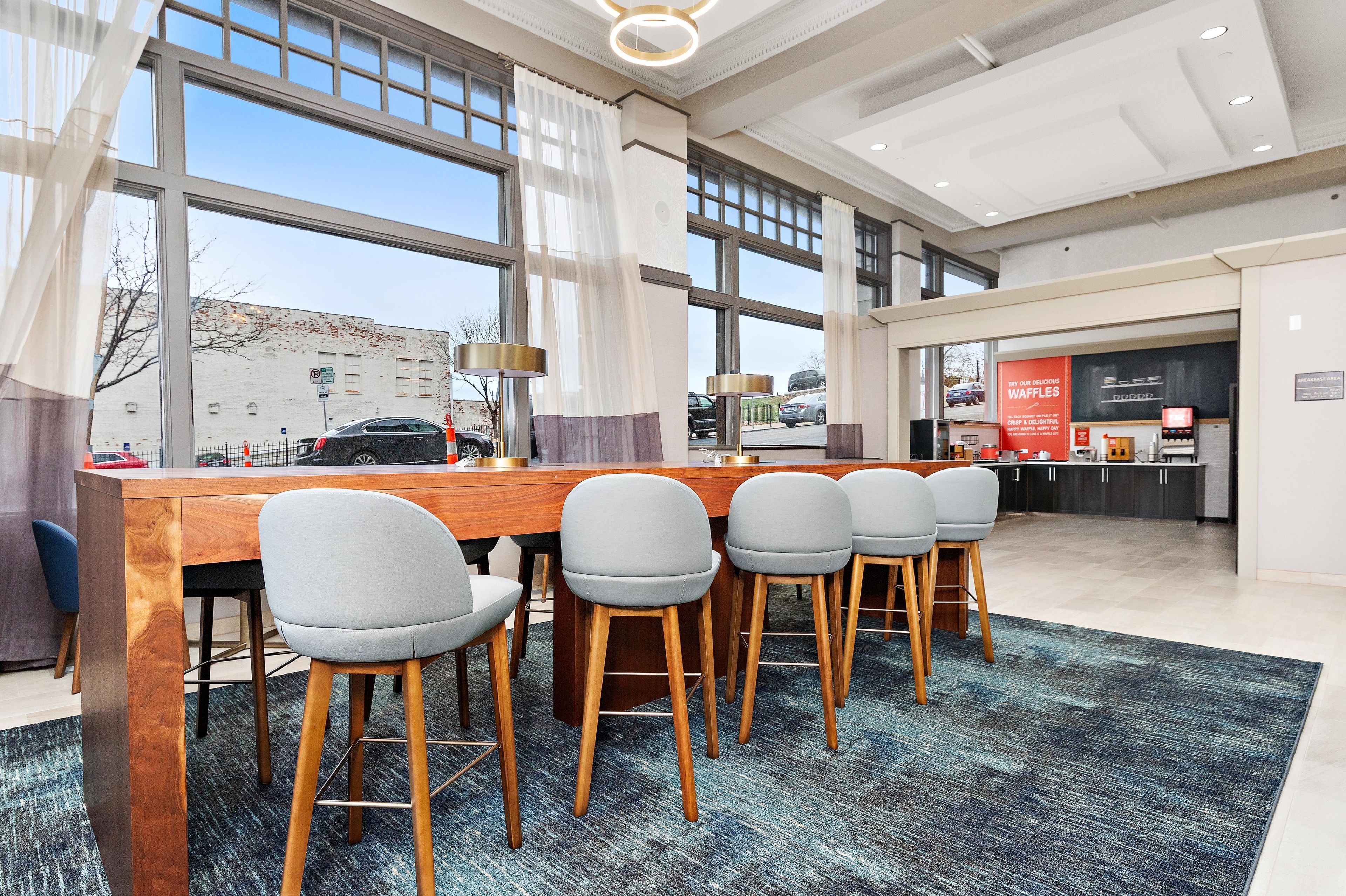 Uma sala de jantar moderna com uma longa mesa de madeira, cinco cadeiras cinzas estofadas e um tapete estampado.
