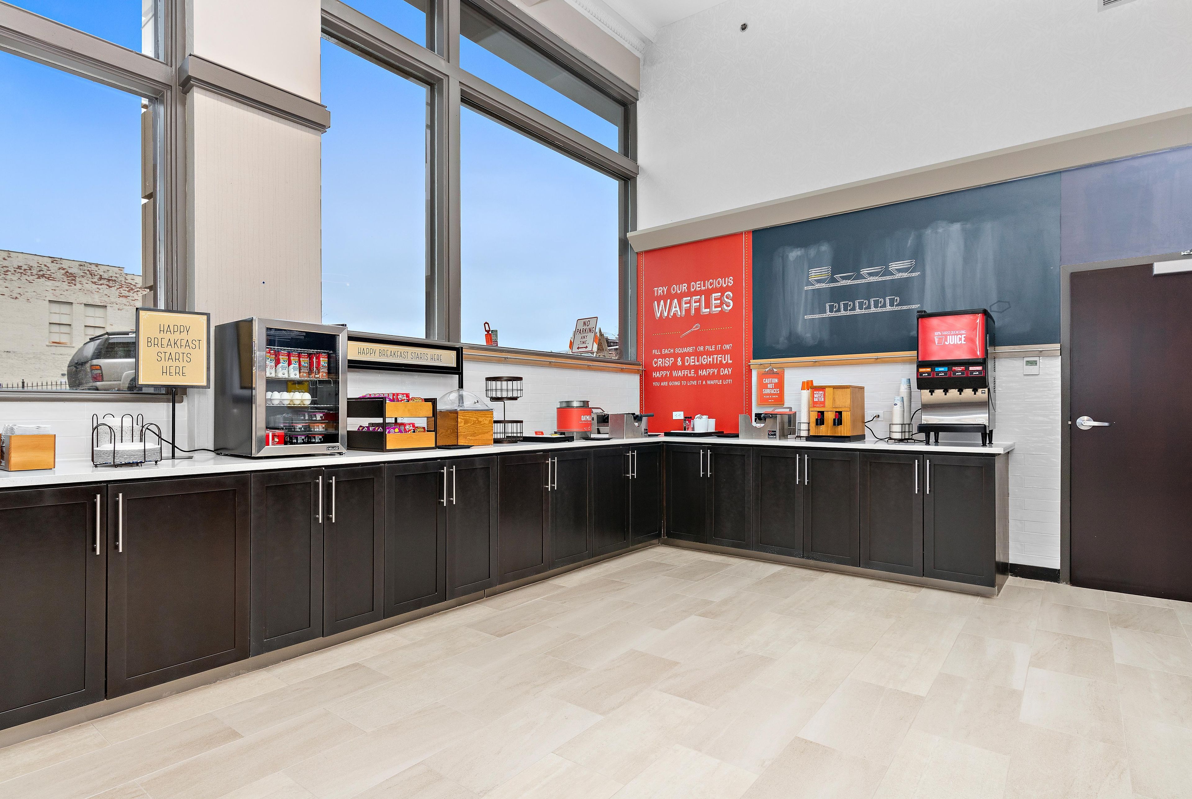 A breakfast bar with waffle and juice dispensers, a chalkboard menu, and a view of the city.