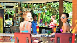 Un espace bar extérieur doté d’un sol carrelé, de chaises rouges et d’un comptoir où sont exposées diverses bouteilles et verres.