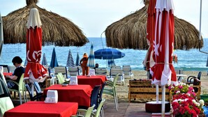 Un restaurant situé en bord de mer, avec une terrasse, des tables recouvertes de nappes rouges et des parasols en chaume.