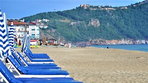 Une plage bordée de rangées de chaises longues et de parasols bleus, une colline rocheuse en arrière-plan et une structure ressemblant à un château au sommet.