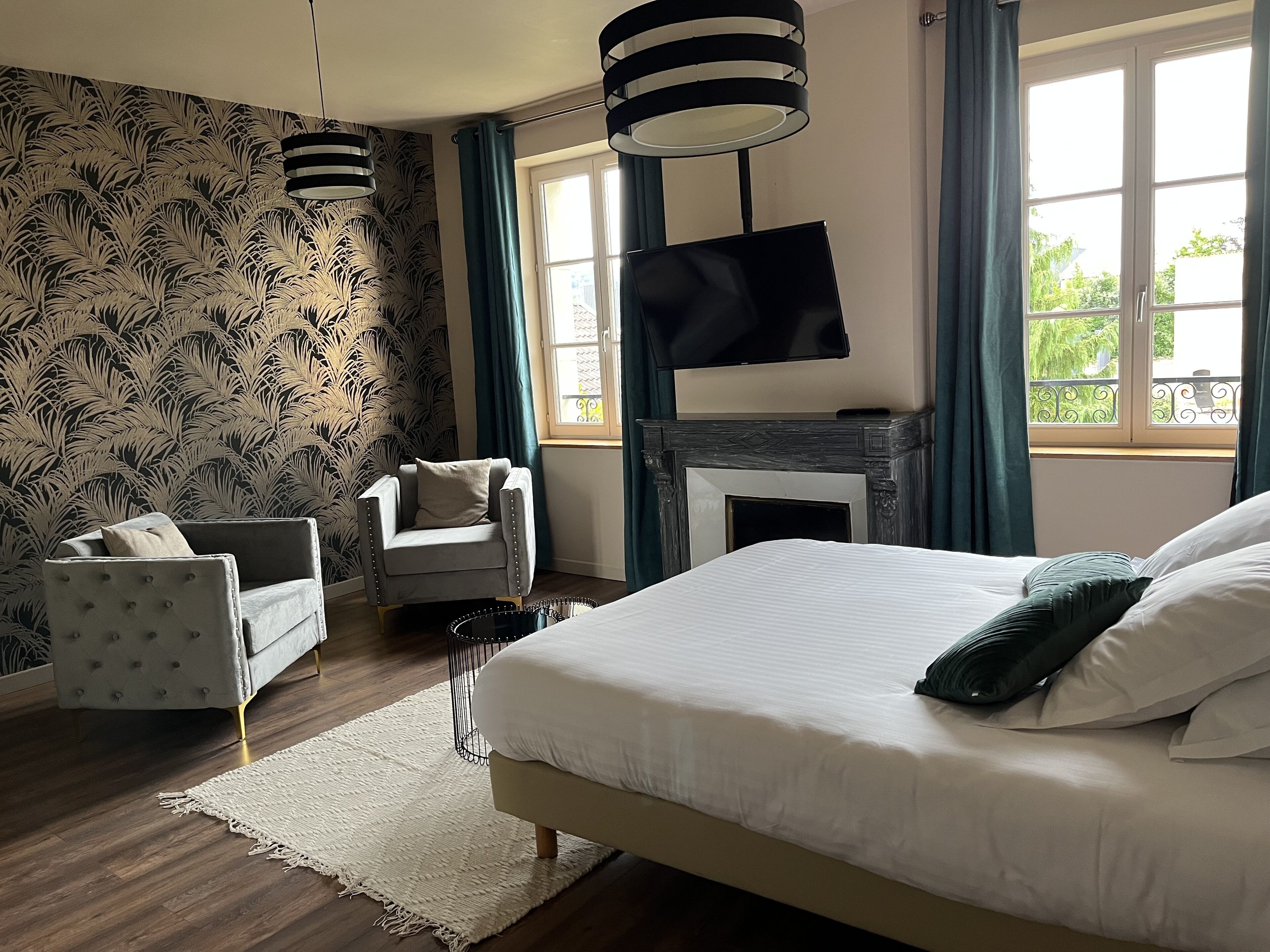 Une chambre à coucher avec un lit, un téléviseur fixé au-dessus d’une cheminée, un fauteuil et un papier peint à motifs.
